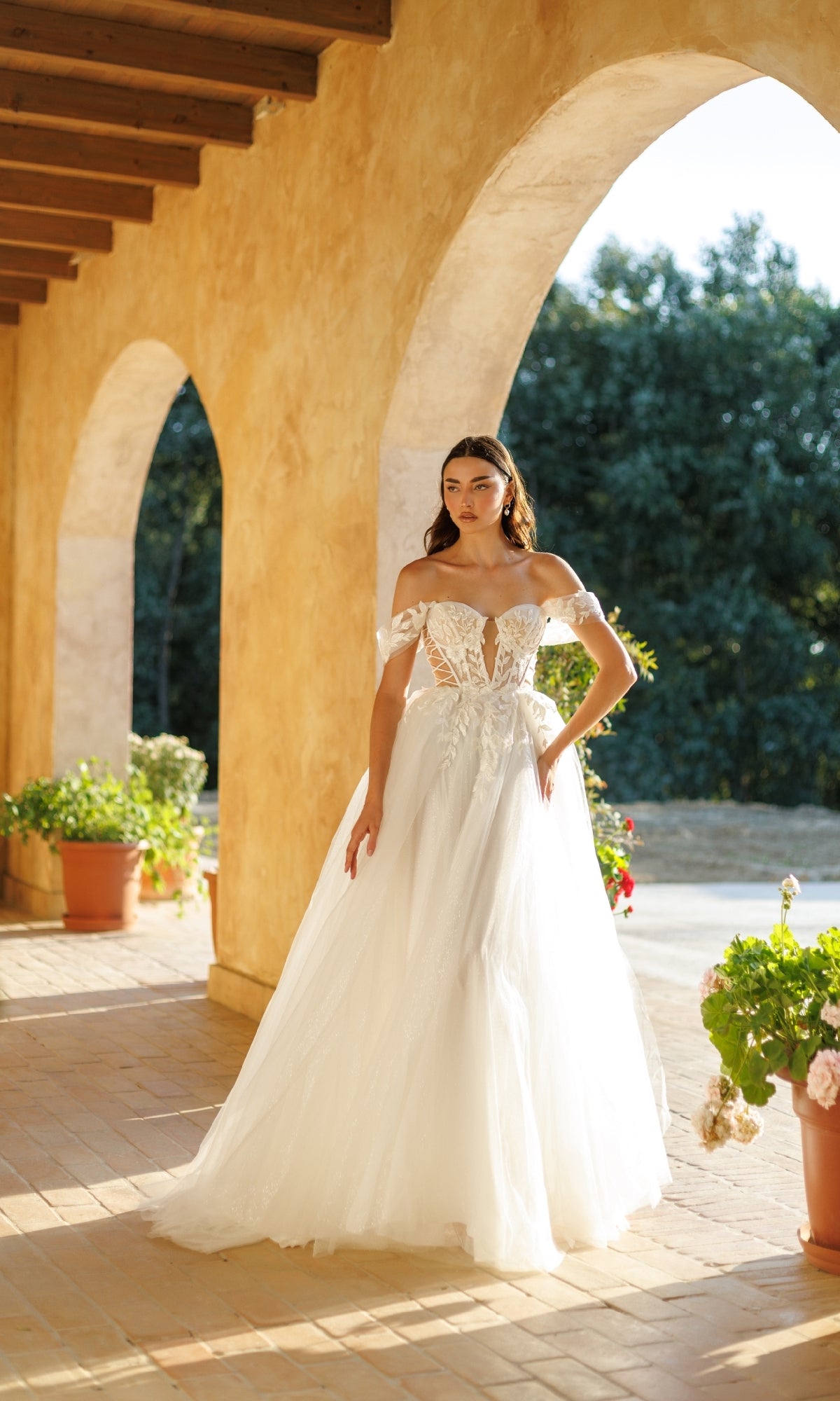 A woman in the Tina Holly TW480 long formal dress, featuring an off-the-shoulder bodice and tulle A-line skirt, stands on a sunlit tiled terrace with potted plants and arched columns.